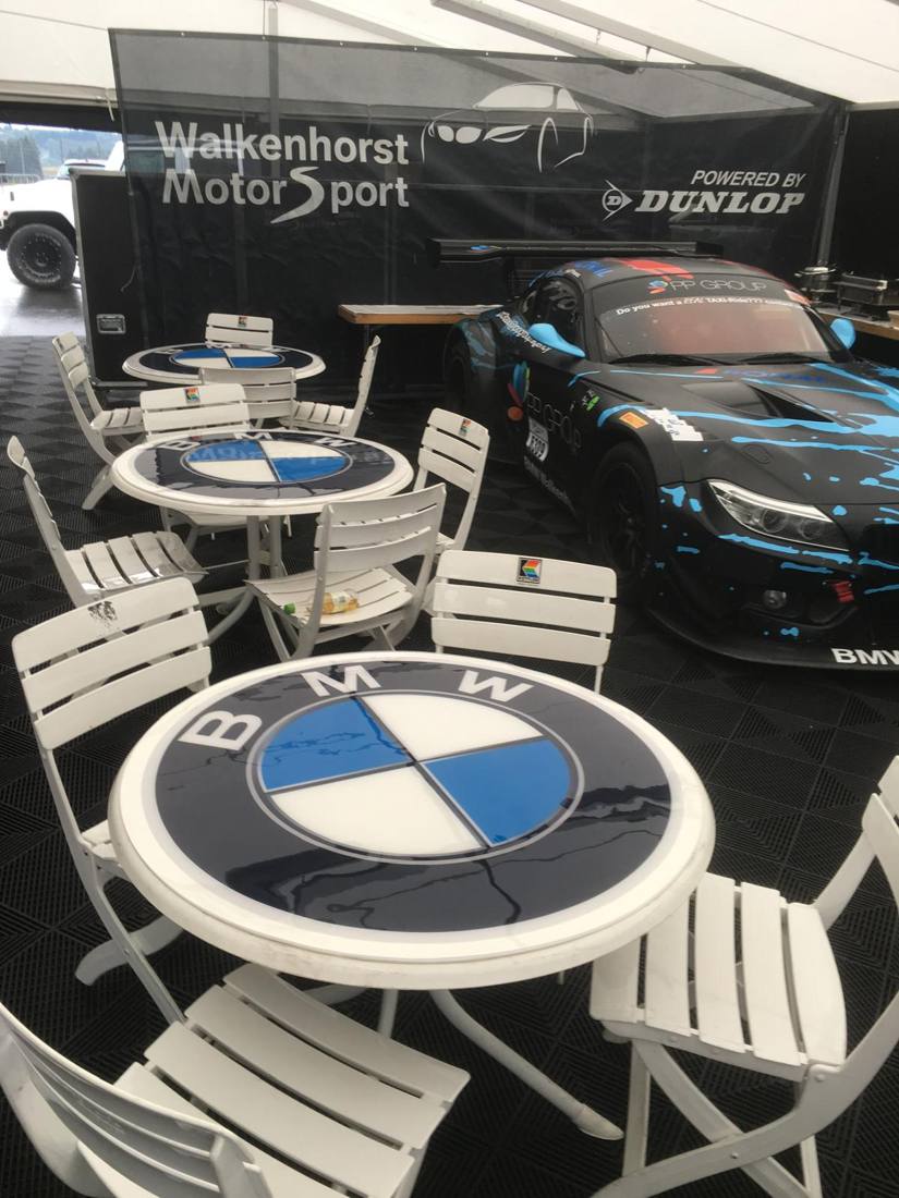 Runde Bistrotische mit Logoauflage vor einem Rennwagen im Hospitality-Zelt