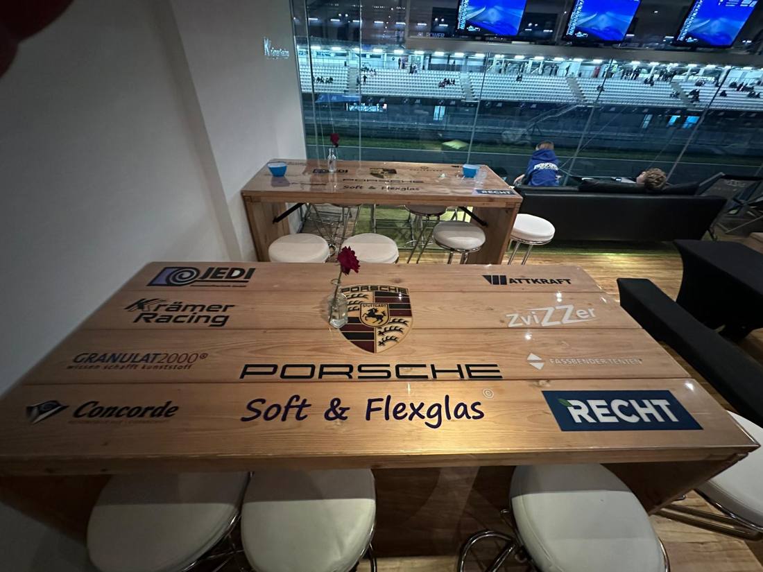 Holz-Stehtische mit Sponsorenlogos unter transparenter Auflage in einer Lounge mit Blick auf die Tribüne